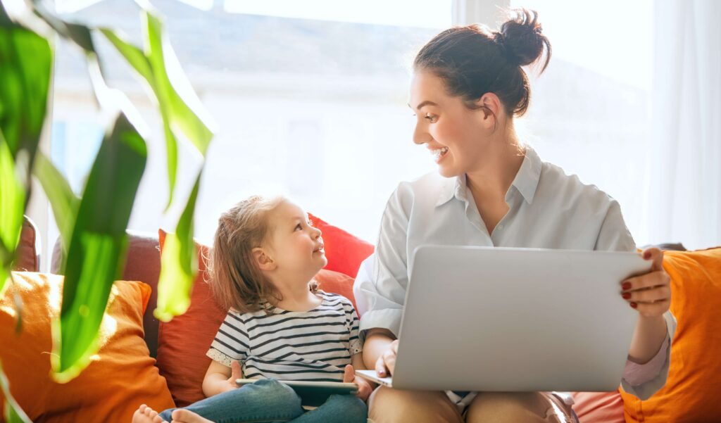 A smiling woman sits on a couch with a laptop on her lap, looking at a young child beside her. The child, holding a tablet, looks up at the woman with a smile. Bright sunlight filters through a window, and colorful pillows and green plants create a warm, cozy atmosphere.