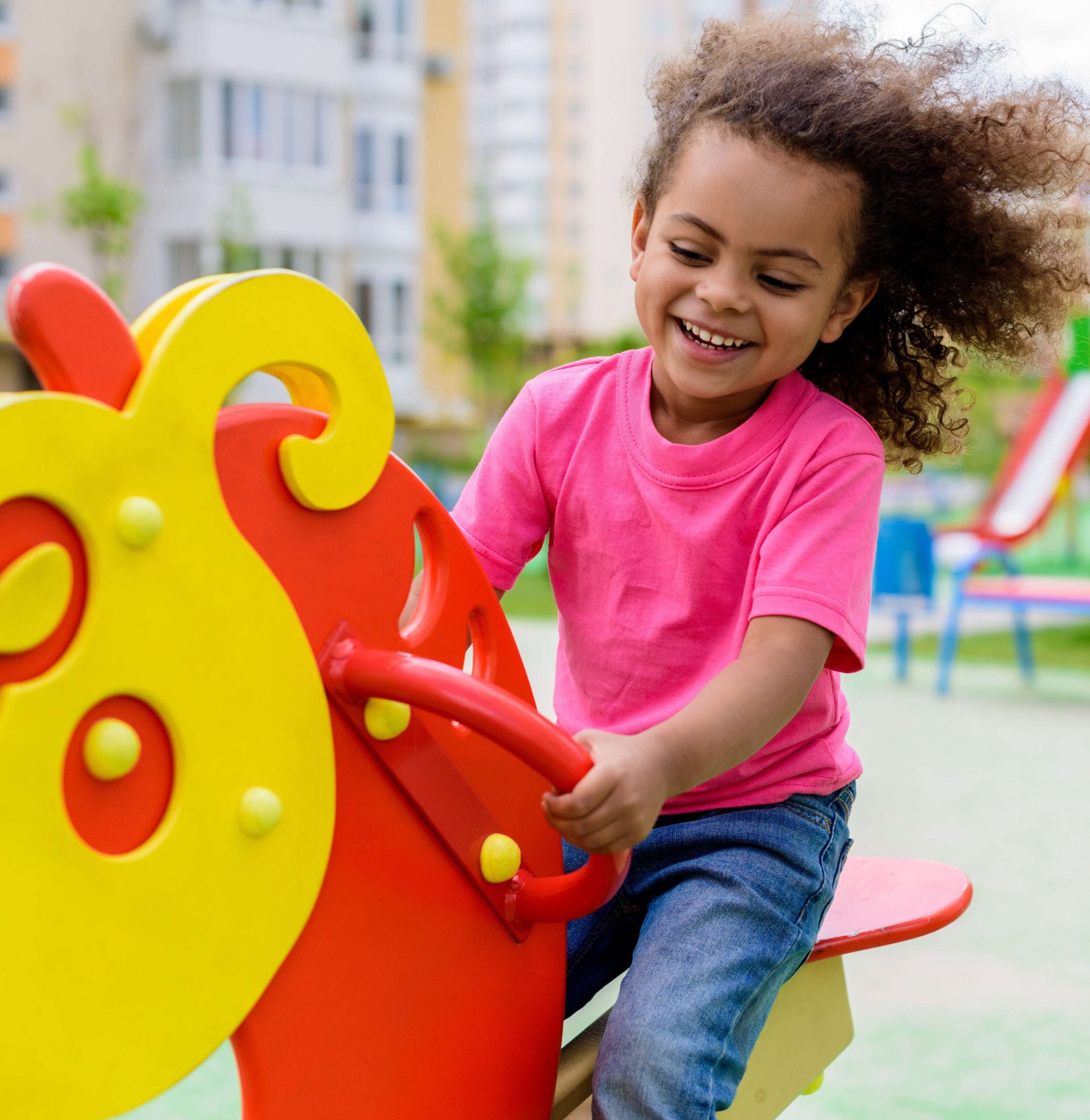 Smiling young child with curly hair wearing a pink shirt and blue jeans, playing on a colorful red and yellow playground rocker outdoors