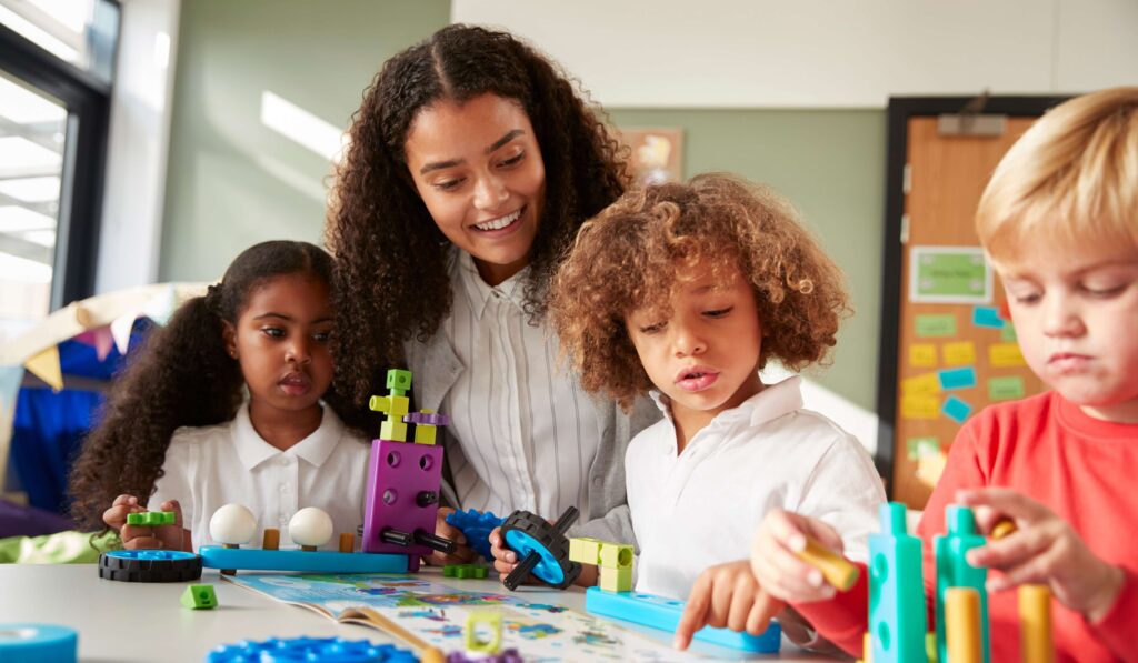 A smiling teacher sits with a group of young children at a table, engaging in hands-on STEM or building activities with colorful blocks and gears. The children are focused and curious, interacting with the materials in a bright, cheerful classroom setting.