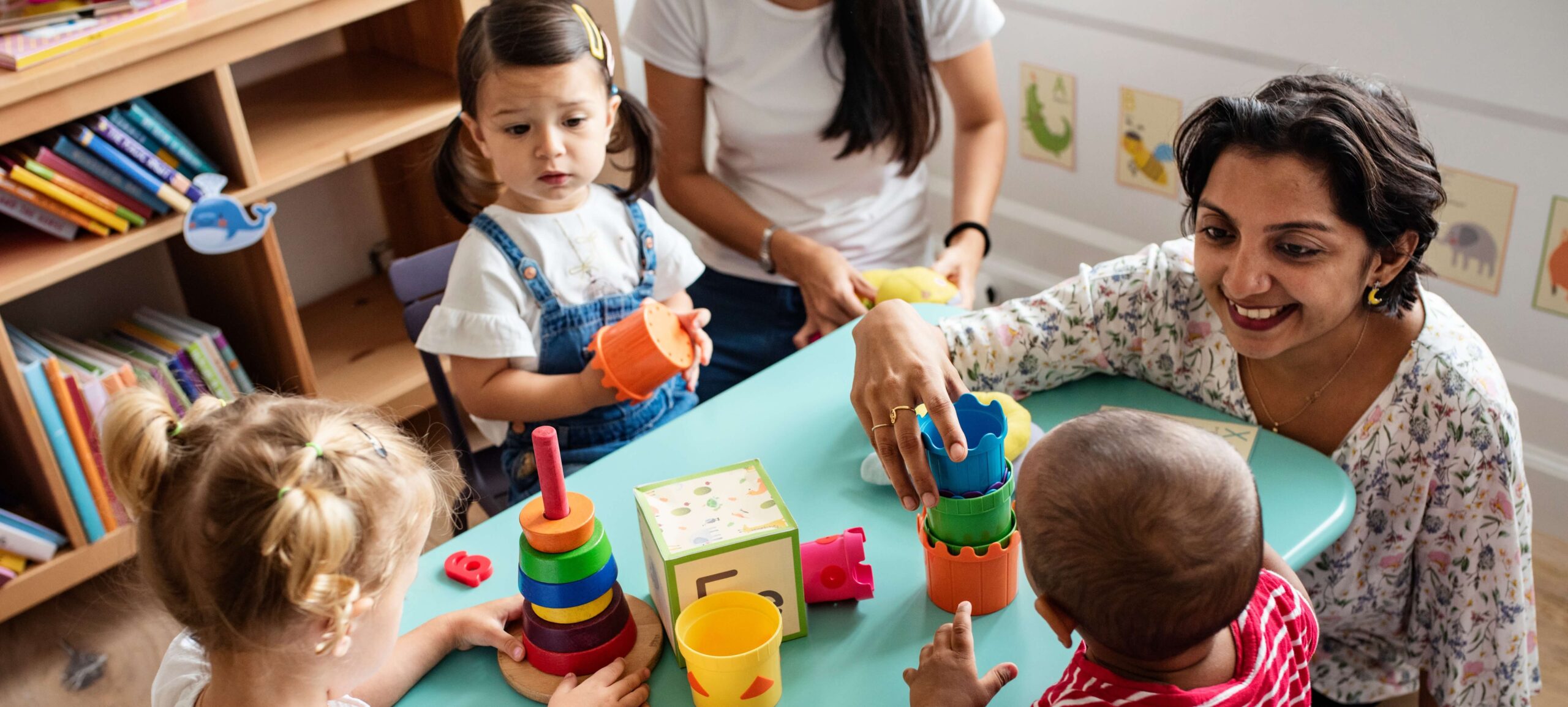 A group of young children and two female caregivers engage in playful learning at a small table in a classroom. Colorful educational toys like stacking cups and shape sorters are spread out on the table. One caregiver smiles warmly at a toddler while the other assists a child holding a stacking toy. Bookshelves and animal wall decor are visible in the background.