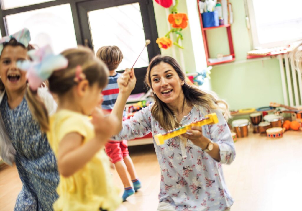 A smiling teacher engaging with young children in a lively classroom, holding a yellow xylophone and a mallet while the children dance and play around her
