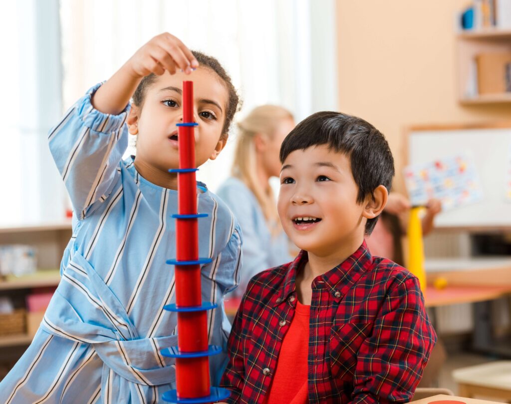 A young girl in a blue striped dress carefully stacks red and blue pieces to build a tall tower, while a smiling boy in a red plaid shirt watches with excitement. They are seated at a table in a bright and cheerful classroom environment.
