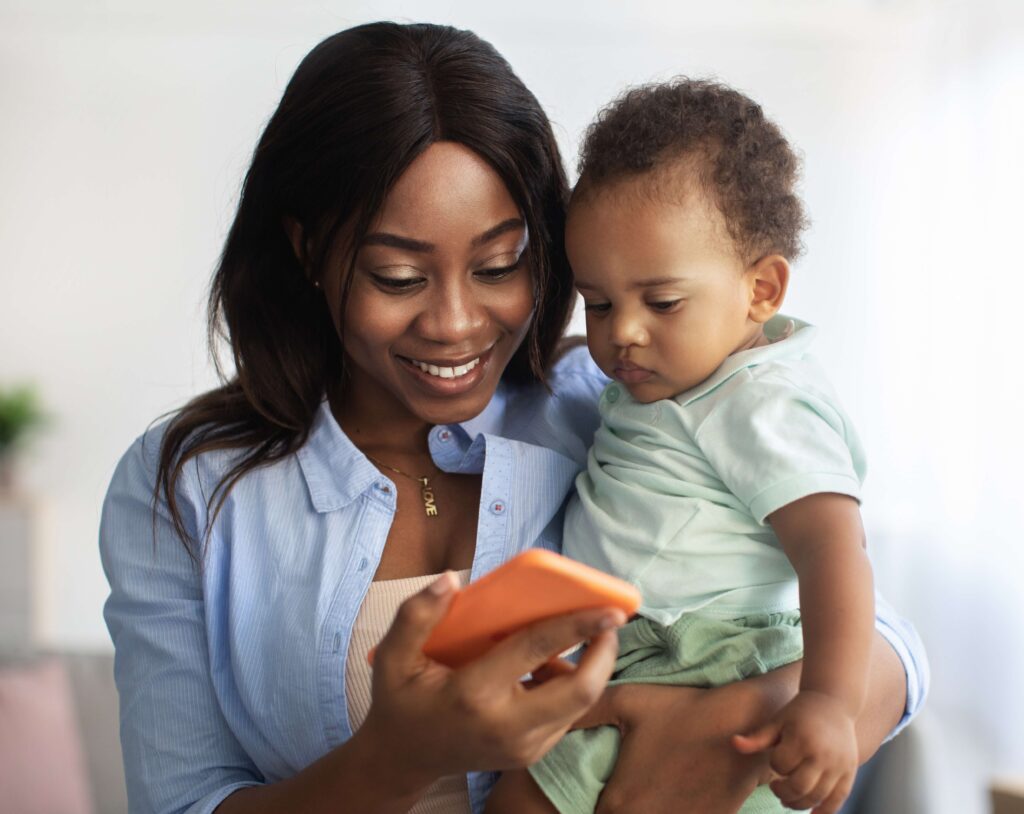 A smiling woman holds a baby in one arm while looking at a smartphone in her other hand. The baby, dressed in a light green outfit, looks at the phone with curiosity. They are indoors in a bright, softly lit setting.