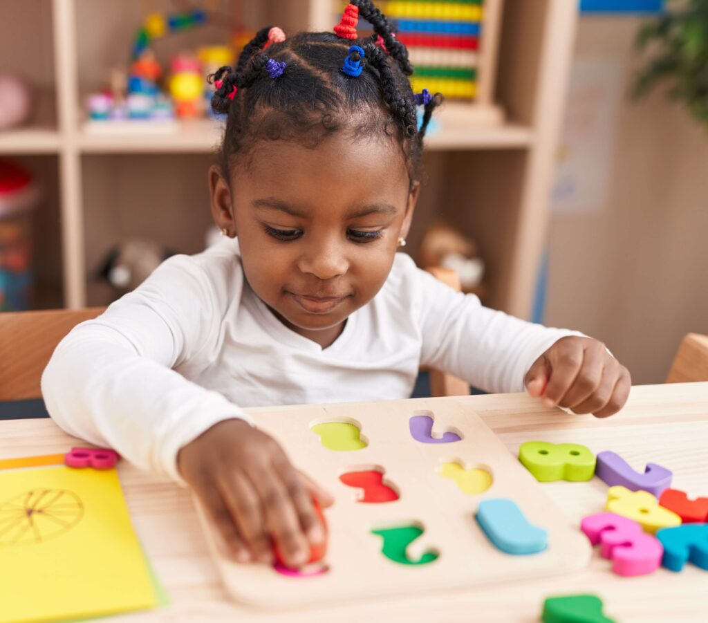 A young girl with colorful braided hair plays with a number puzzle at a table in a classroom. She is focused as she places a red puzzle piece into its matching slot. Bright educational toys and shelves are visible in the background.