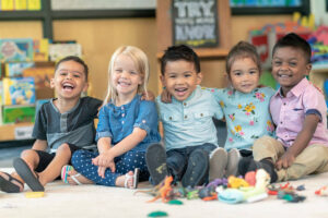 Five young children of diverse backgrounds sitting together on the floor in a classroom, smiling and with arms around each other, with toys scattered in front of them