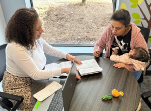 Two women sitting at a table reviewing documents, one holding a pen and a baby, the other pointing at the papers, with a laptop, toy car, and peeled orange on the table