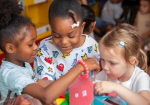 Three young children playing together with colorful toys in a classroom, smiling and focused on their activity