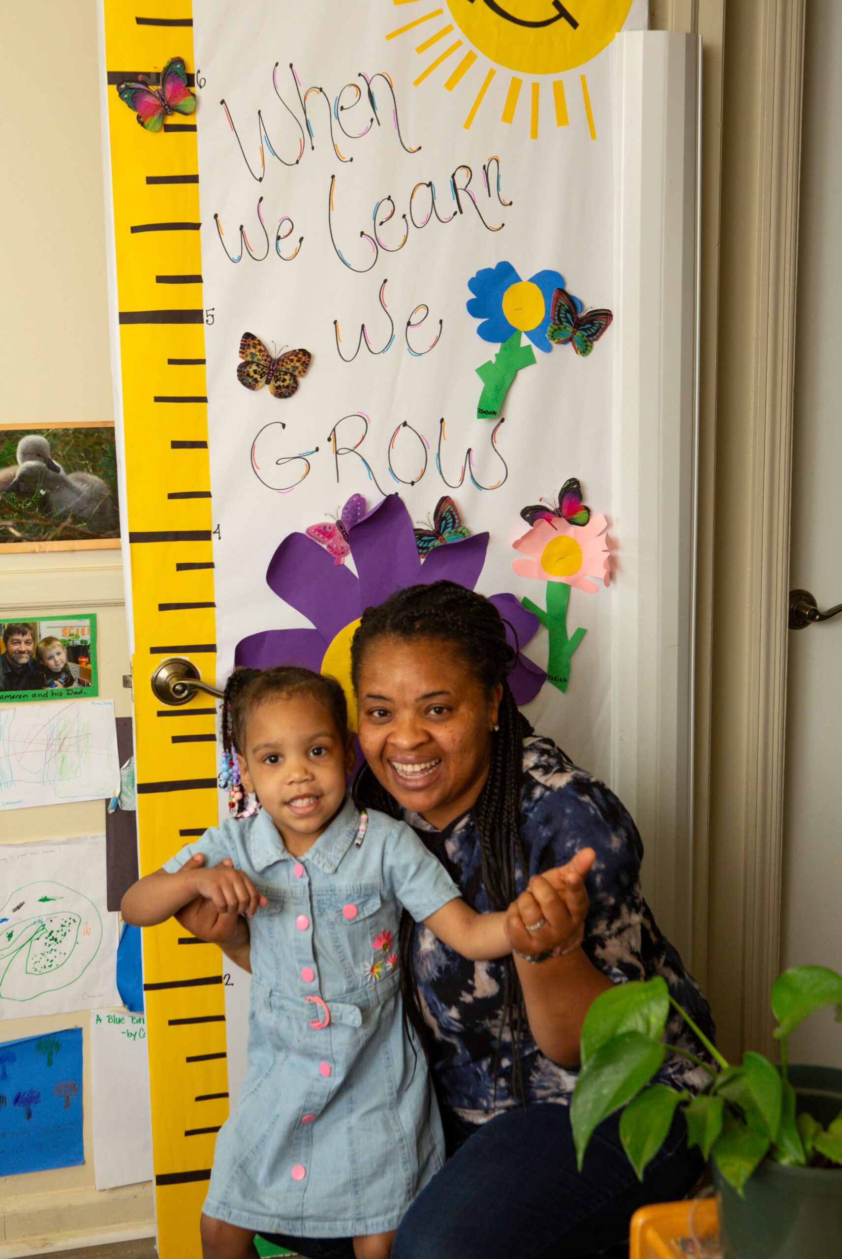 A smiling woman and a young girl stand together in front of a colorful classroom height chart decorated with butterflies, flowers, and a sun. The chart features the handwritten phrase “When we learn we grow.” The girl wears a light blue denim dress with pink buttons, while the woman wears a blue patterned shirt. Artwork and photos are displayed on the wall beside them, and a green potted plant is in the foreground