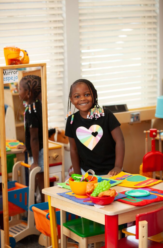 A smiling young girl with braided hair decorated with colorful beads stands in a play kitchen area. She is wearing a black t-shirt with a pastel rainbow heart design and denim shorts. In front of her is a small table set with colorful placemats, bowls, and toy food including grapes and bread. A mirror behind her reflects her image, and the word “Mirror / espejo” is labeled at the top. Brightly colored chairs, a play shopping cart, and other toys are visible in the background