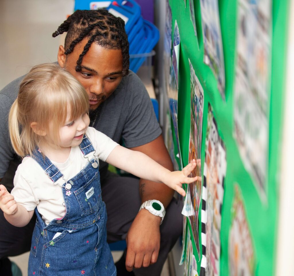 A young girl wearing denim overalls points at photos on a green bulletin board while smiling. A man crouches beside her, attentively looking at the display and engaging with her. The setting appears to be a classroom or learning environment.