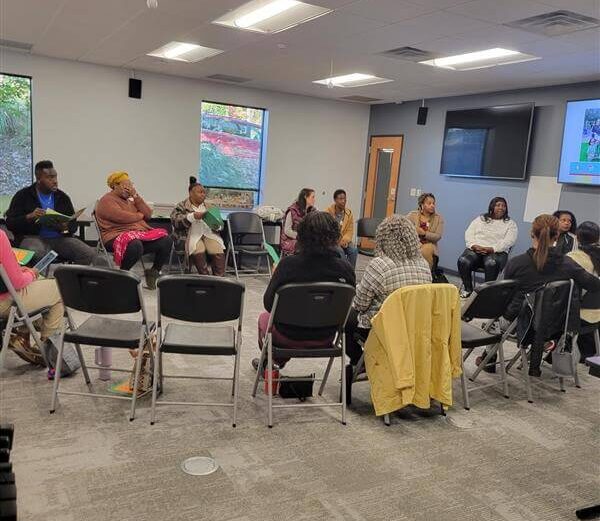 A group of adults sit in a circle of chairs during a professional development session in a classroom.