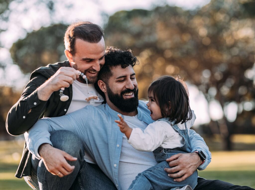 Two smiling men outdoors holding and playing with a young child, one man showing the child a small toy while the other holds the child on his lap