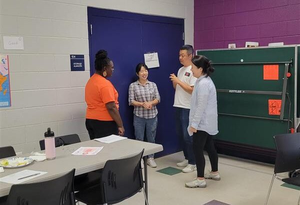 Four adults stand in a casual group conversation near a blue door in a classroom setting. One woman wears an orange shirt and faces three others who are smiling and engaged. Behind them is a green folded ping-pong table and a purple accent wall. Nearby tables have papers, plates, and a water bottle.