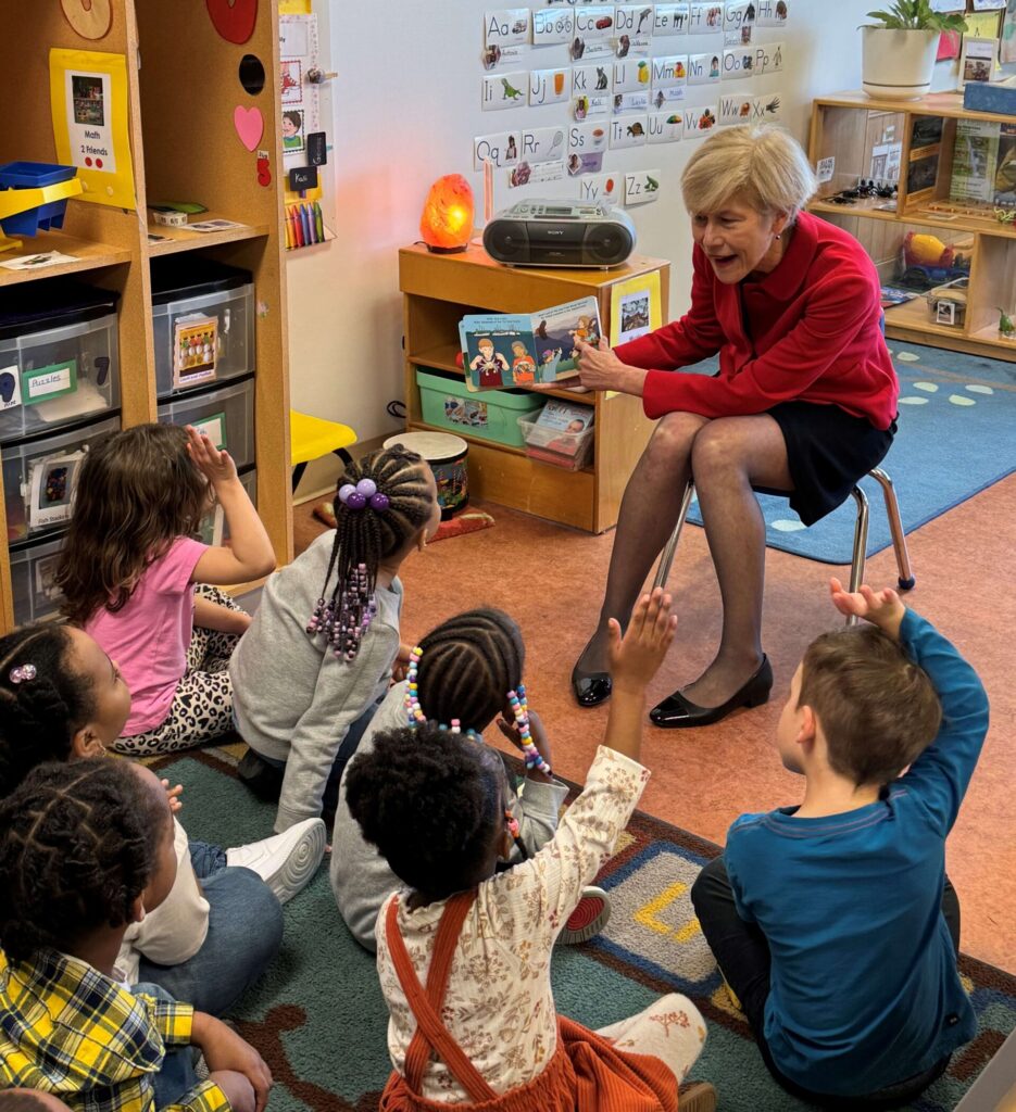 A woman in a red sweater reads a picture book to a group of young children sitting on the classroom carpet. Several children raise their hands eagerly to answer questions. Behind her is a “Letter Wall” with the alphabet displayed, along with classroom storage and decorations.