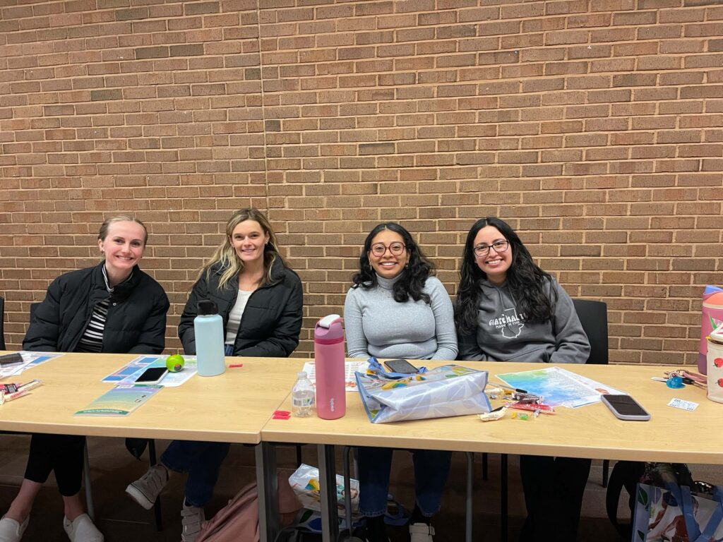Four women sitting at a table in front of a brick wall, smiling at the camera. The table is covered with water bottles, papers, phones, and various snacks and supplies.