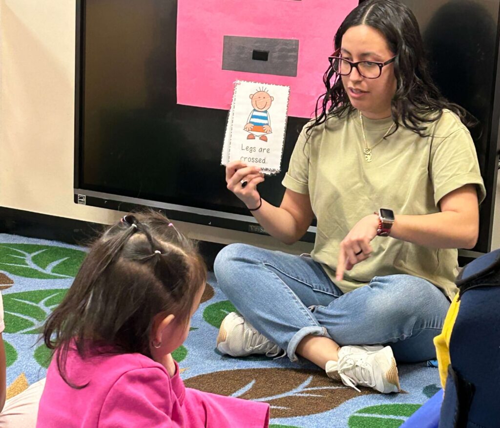 A teacher shows a child a visual card with the words “Legs are crossed” while sitting together on the classroom carpet.