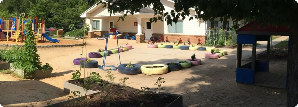 Outdoor playground and garden area with play structures, painted tire planters, and tricycles near a school or childcare building.